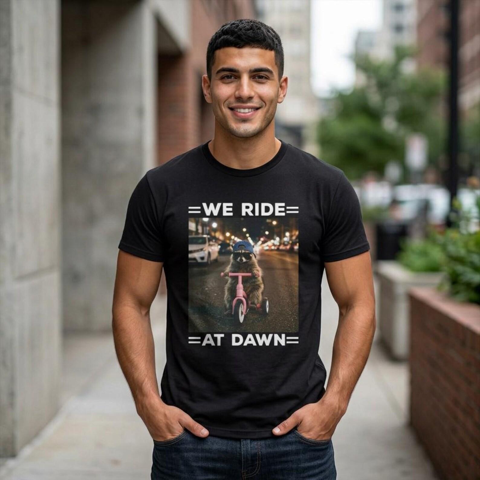 We Ride at Dawn Funny Raccoon T-Shirt; Raccoon Riding Tricycle Chaotic Meme Tee; Absurd Animal Humor Graphic Top S