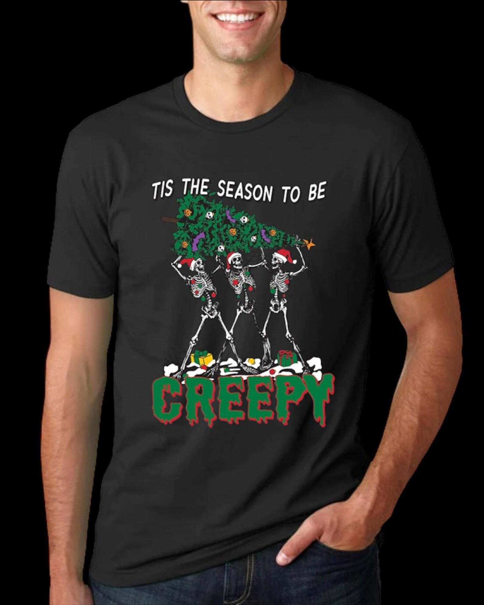 Get into the spooky holiday spirit with this fun “‘Tis the Season to be Creepy” T shirt! Featuring three dancing skeletons hauling a decorat M